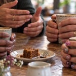 Women with different nail styles, from bare nails to colourful nail art, hold cups around a café table.